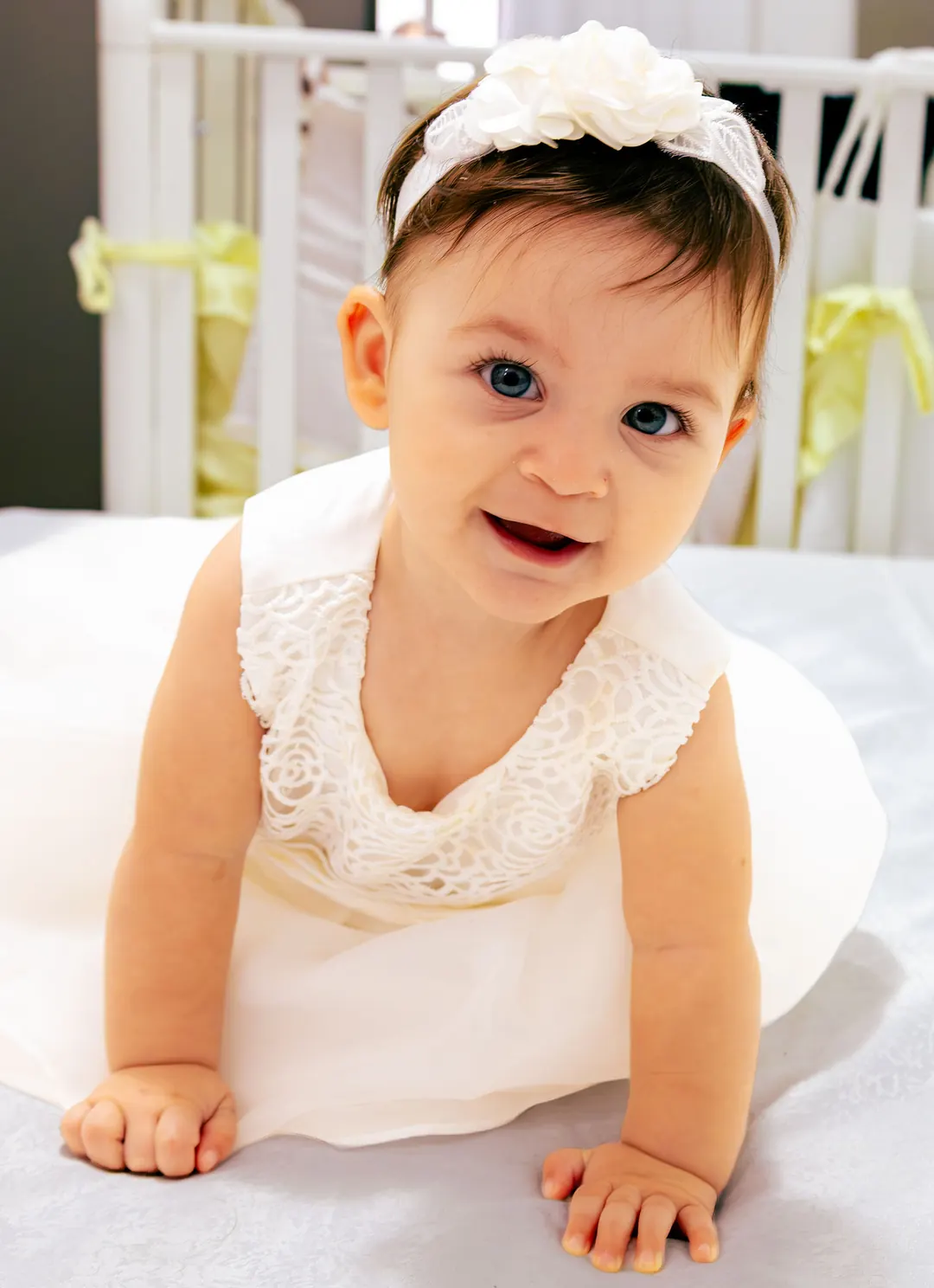 Una bimba con il vestito bianco e la fascia di fiori sorride gattonando.