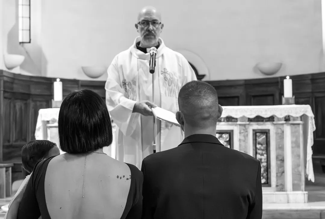 In bianco e nero, il sacerdote all'altare durante la cerimonia, con i genitori di spalle.