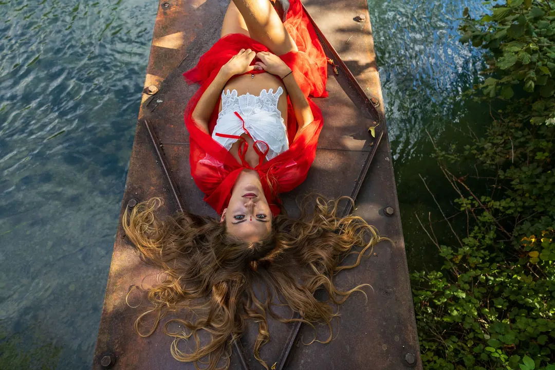 Una ragazza con il mantello rosso sdraiata su un pontile di legno sopra l'acqua, vista dall'alto.