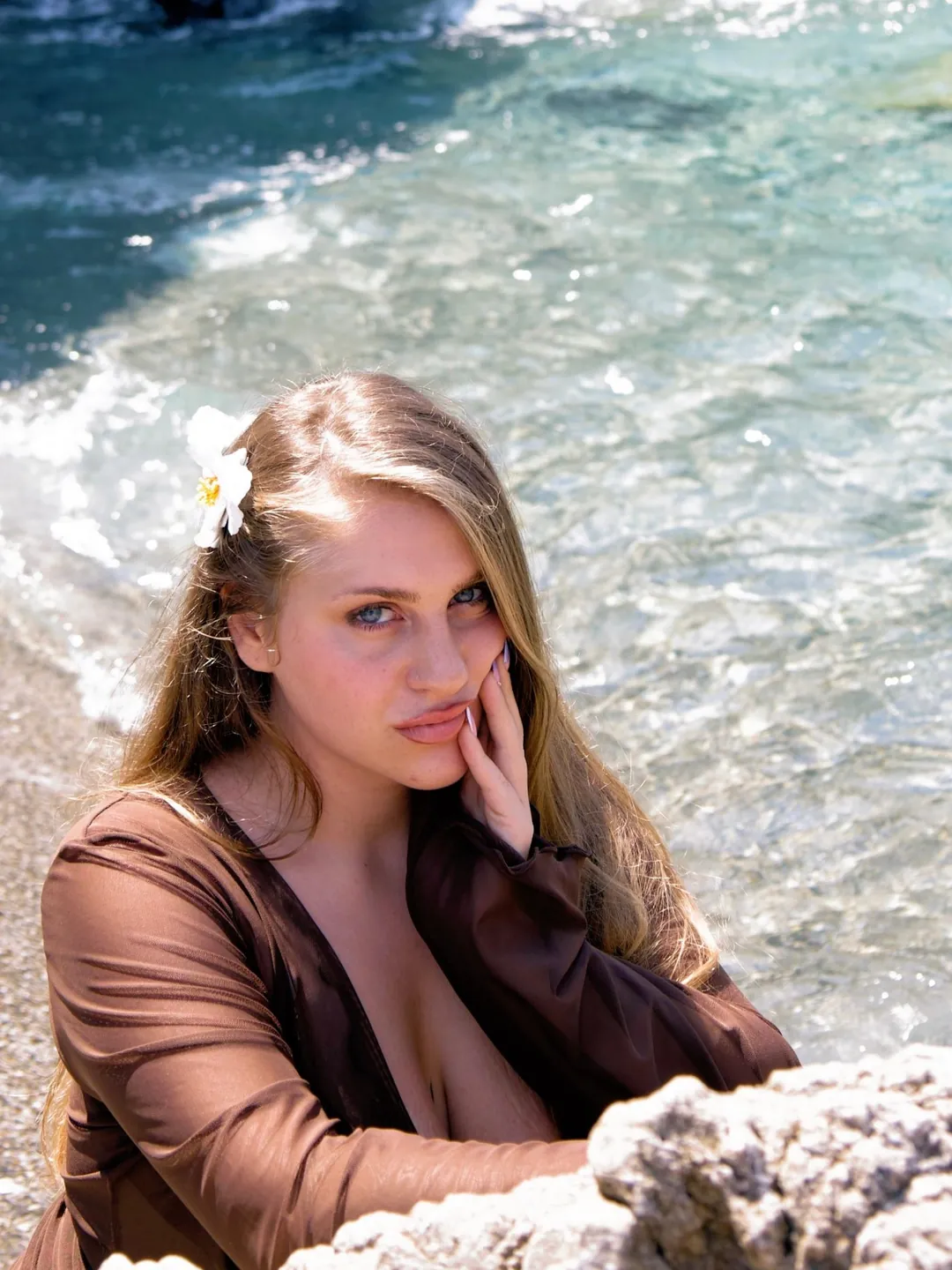 Una ragazza con un fiore bianco tra i capelli, appoggiata agli scogli, con il mare turchese alle spalle.